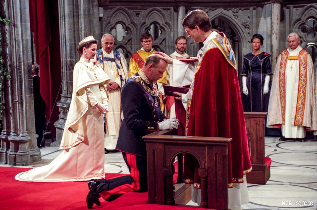 Kong Harald står på kne og blir signet av en biskop. I bakgrunnen står dronning Sonja og flere biskoper. Foto.
