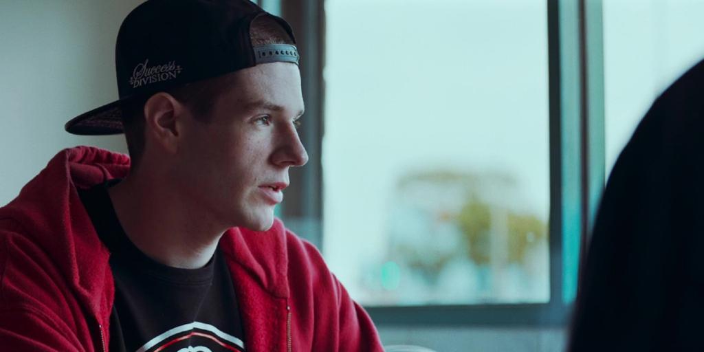 Photo of the actor Connor Swindells as the character Adam in the film VS. A teenage boy in a black cap, black t-shirt and a red hoodie. 