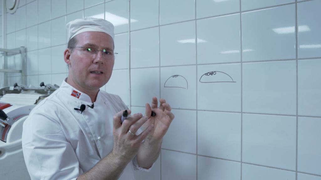 En mann i bakeklede teiknar med tusj på flisene på veggen mens han gestikulerer med hendene. Han har teikna to brød. Foto.