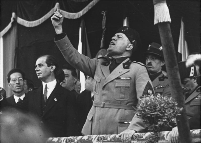 Mussolini i uniform står på en scene, heiser hånden og taler til massene. Foto.
