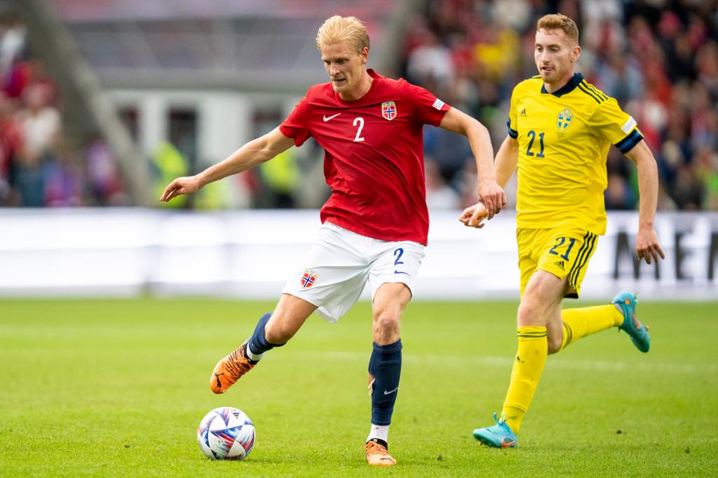 Fotballspelar med norsk landslagsdrakt i raudt og kvitt har ballen, mens ein svensk spelar i gul drakt spring bak han. Dei ser begge egga ut midt i kampens hete. Foto.