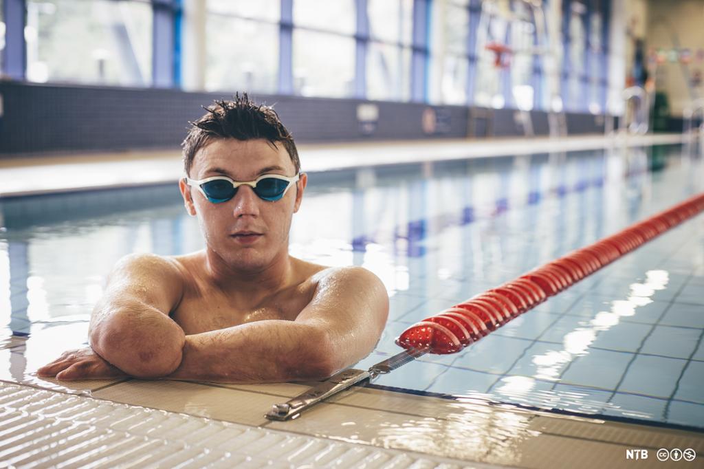 En mann med en amputert arm er i et svømmebasseng og holder seg i kanten med armene. Han har svømmebriller på. Foto.