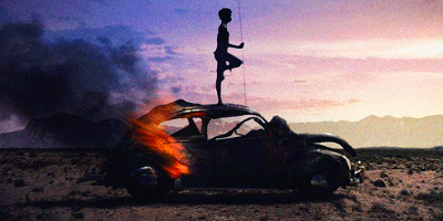 Silhuett av gut som står på toppen av ein brennande bil. Tørt og aude landskap i bakgrunnen.