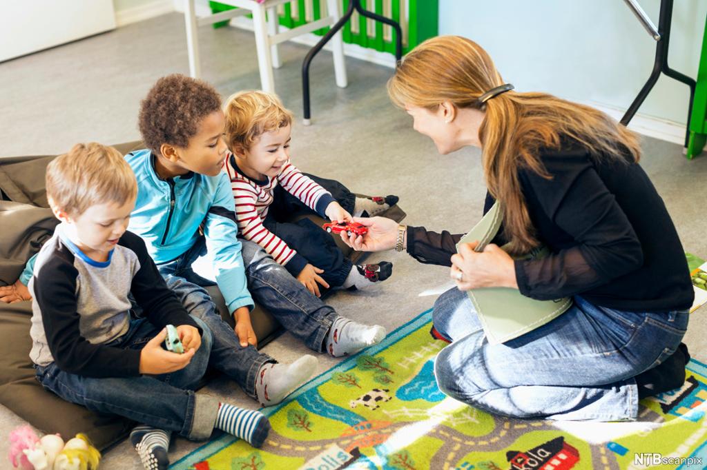 En voksen og tre barn sitter på gulvet. Den voksne rekker en lekebil til det ene barnet. Foto.