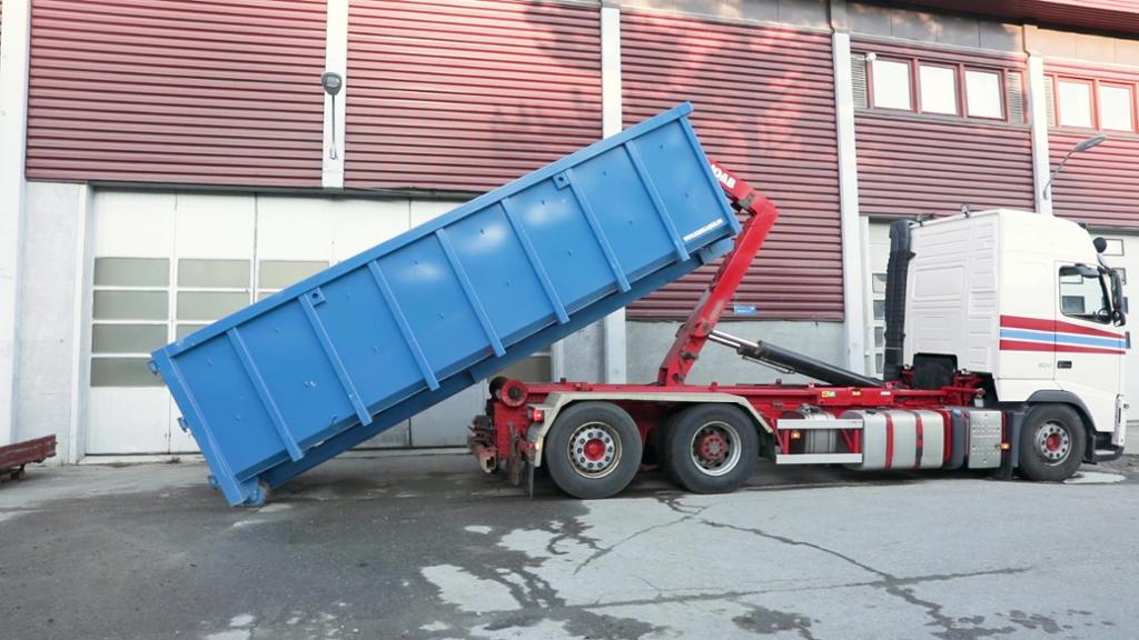 En krokbil står parkert utenfor et lagerbygg og har tippet en stor, blå konteiner i nesten 45 graders vinkel. Foto.
