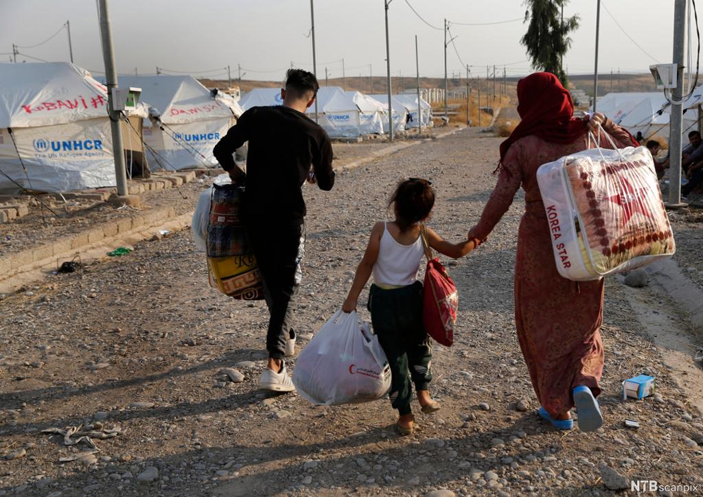 En liten syrisk familie med tepper  og poser på vei gjennom en flyktningeleir i Irak med telt fra FN. Foto. 
