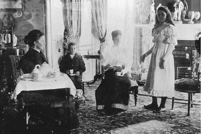 Svart-hvitt foto av pent kledde mennesker i ei fin stue. To kvinner og en gutt sitter med tekopper i hendene, og ei ung kvinne står. Foto. 