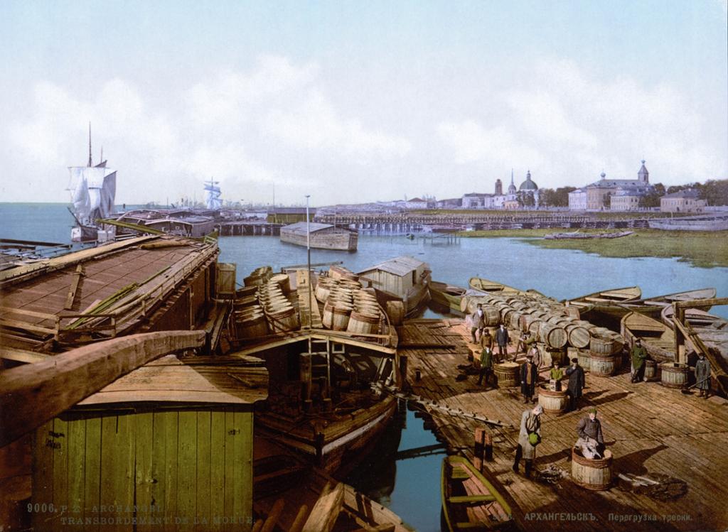 Havna i Arkhangelsk i 1896. Båter lastet med tønner i forgrunnen, større skip og en del bebyggelse i bakgrunnen. Noen mennesker jobber på ei brygge i forgrunnen. Foto.