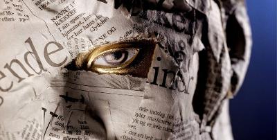 Ansikt fullklistret med biter av avispapir. Bare et åpent øye er synlig. All synlig hud rundt øyet er malt i gull. Fotografi.