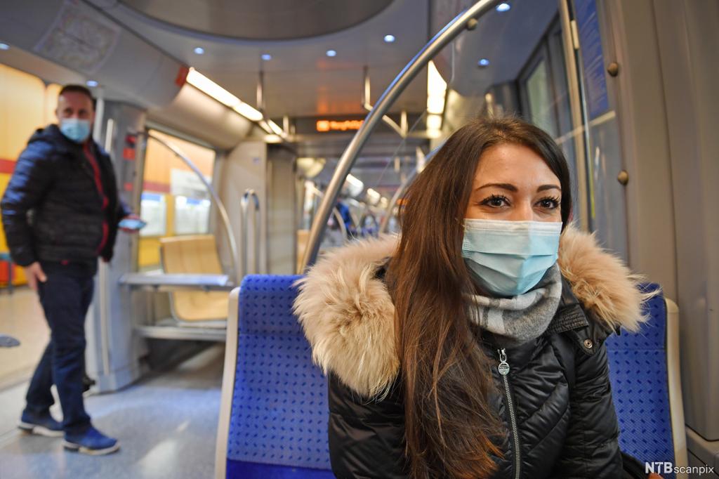 Ung kvinne sitter på T-banen med munnbind på. Mannlig passasjer i bakgrunnen har også munnbind. Foto. 