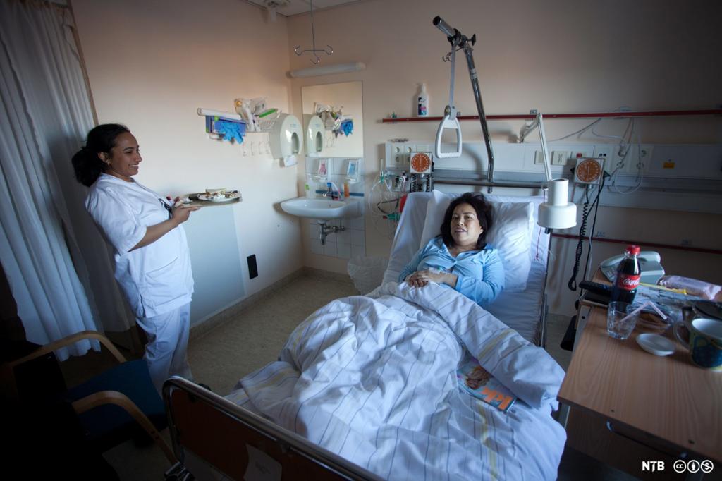 En sykepleier står med et brett med mat ved siden av senga til en kvinnelig pasient på et sykehusrom. Pasienten ligger halvveis sittende. Foto.