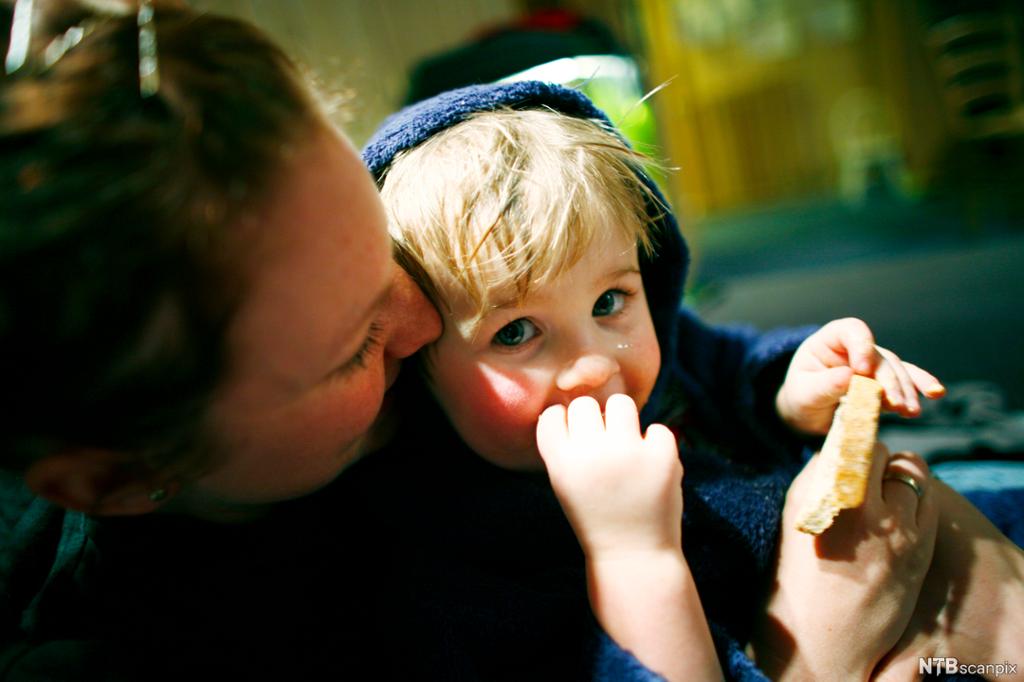 Et lite barn sitter på fanget til en kvinne. Kvinnen holder ei brødskive i hånden, barnet plukker på brødskiva og tar den ene hånden til munnen. Foto. 