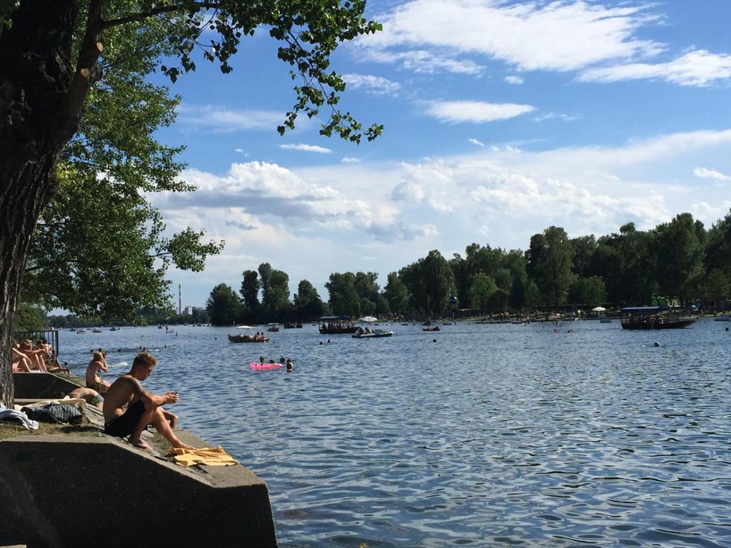 Badestrand ved Donau, med folk som soler seg. Småbåter på vannet og trær i bakgrunnen. Foto.