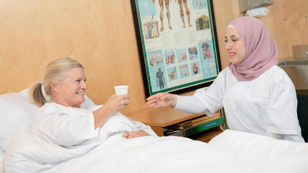Kvinnelig pasient får servert en kopp drikke i senga av en kvinnelig helsefagarbeider. Foto.