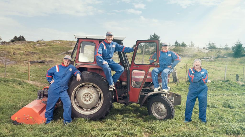 Fire blide personar i blå kjeledressar sit på eller står ved sida av ein gammal traktor på ei eng. Foto.