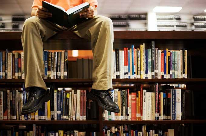 Mann som sitter på toppen av en bokhylle og leser i ei bok. Foto. 