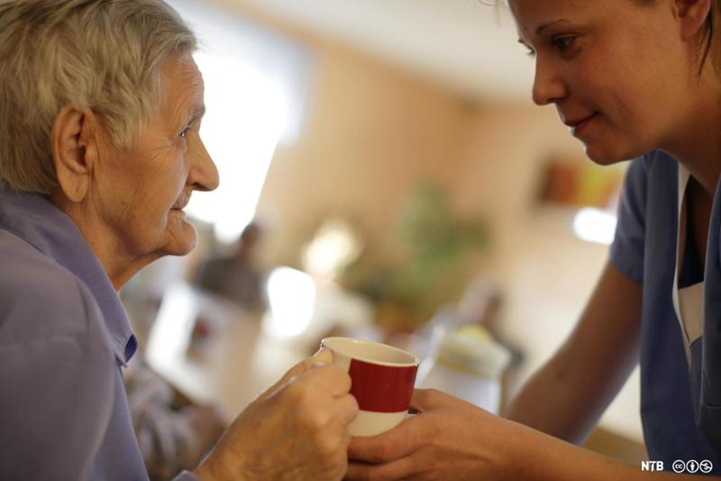 Helsefagarbeider hjelper gammel dame på sykehjem med å holde kaffekoppen. Foto.
