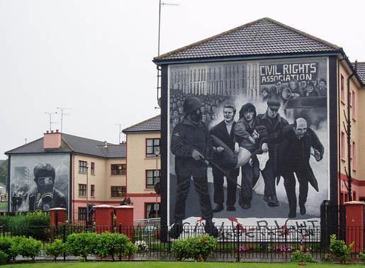 Photo: Mural commemorating Bloody Sunday. We see the words 'Civil Rights Association'. A soldier. People carrying a bleeding victim. Protesters in the background. 
