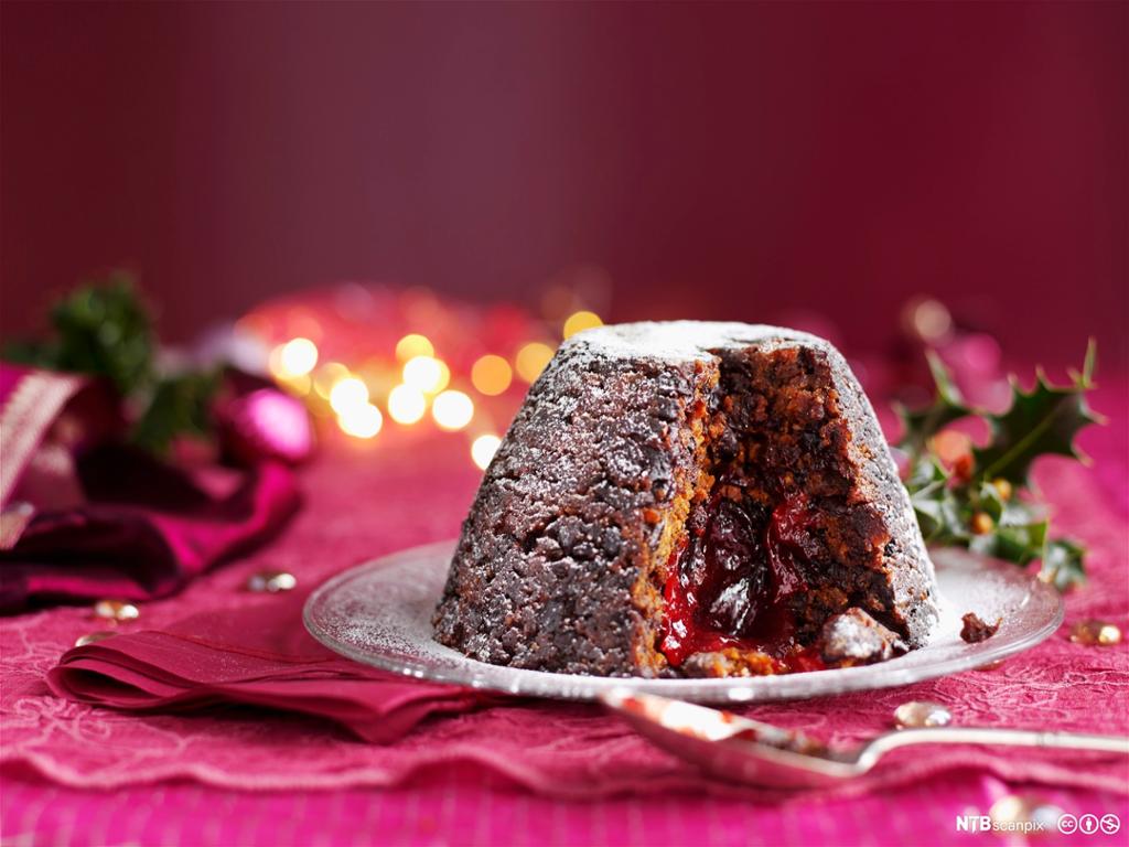 Eit bord med rosa duk og ein glastallerken med julepudding som det er teke eit stykke av. Foto.