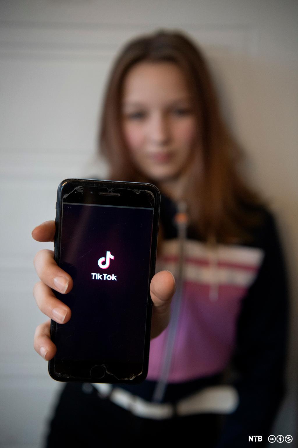 Ei jente viser fram mobilen til kamera. På skjermen står logoen til appen TikTok, et populært sosialt medium på 2020-tallet. Foto.