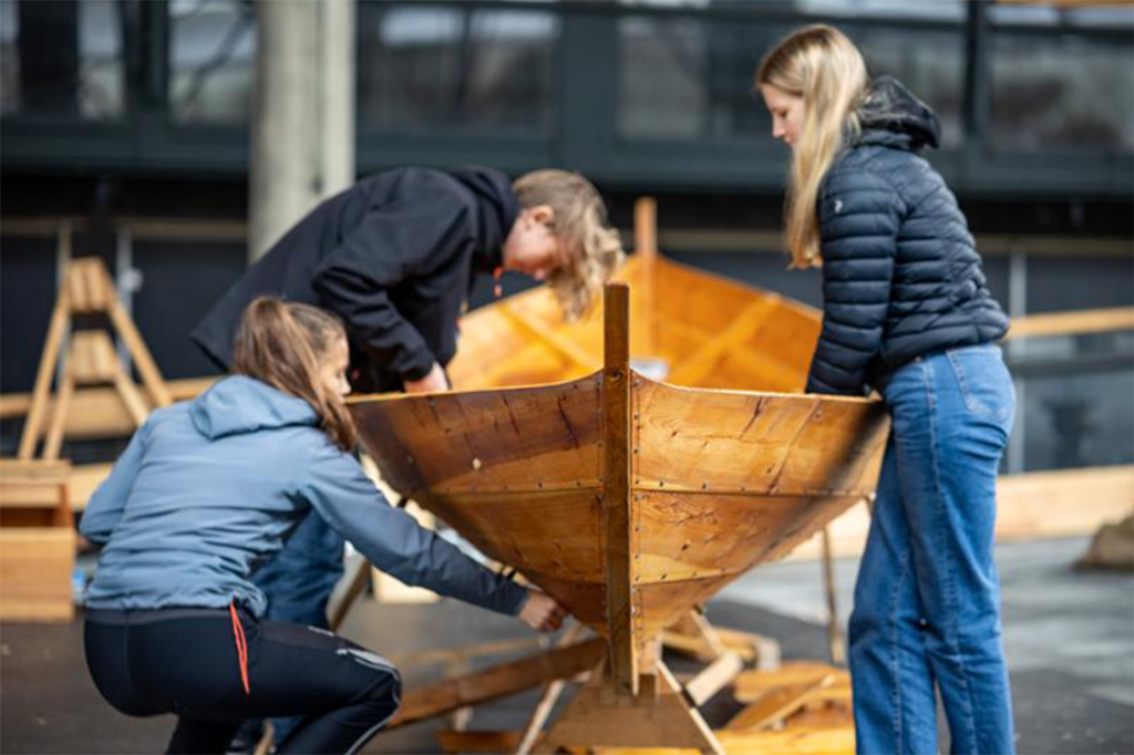 Tre elever arbeider på en liten trebåt. Foto. 
