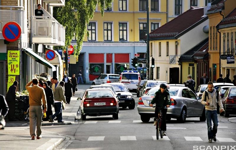  En gate med butikker, biler og gående og syklende personer. Foto.