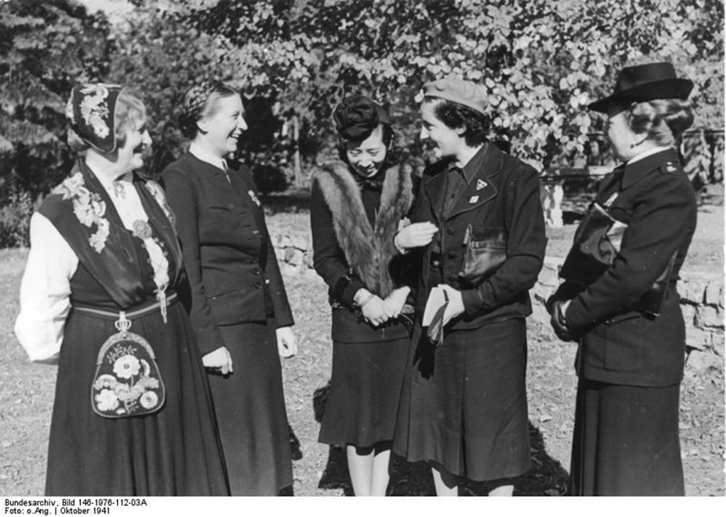Fem kvinner står og snakker sammen. En er kledd i bunad, de andre i drakt og hatt. Historisk foto.