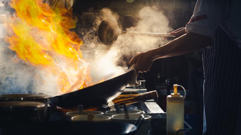 En kokk står ved en komfyr og rører i en wok med store flammer. Foto.