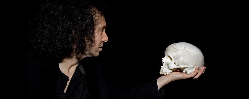 A photo of Hamlet holding a white skull. The actor is dressed in black and he's standing in front of a black background.