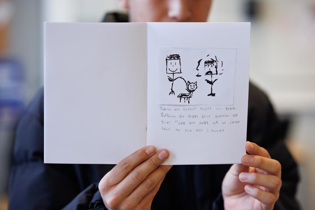 En person holder opp ei kladdebok med tegning av to strekmenn og en katt. Det er håndskrevet tekst under bildet. Foto. 