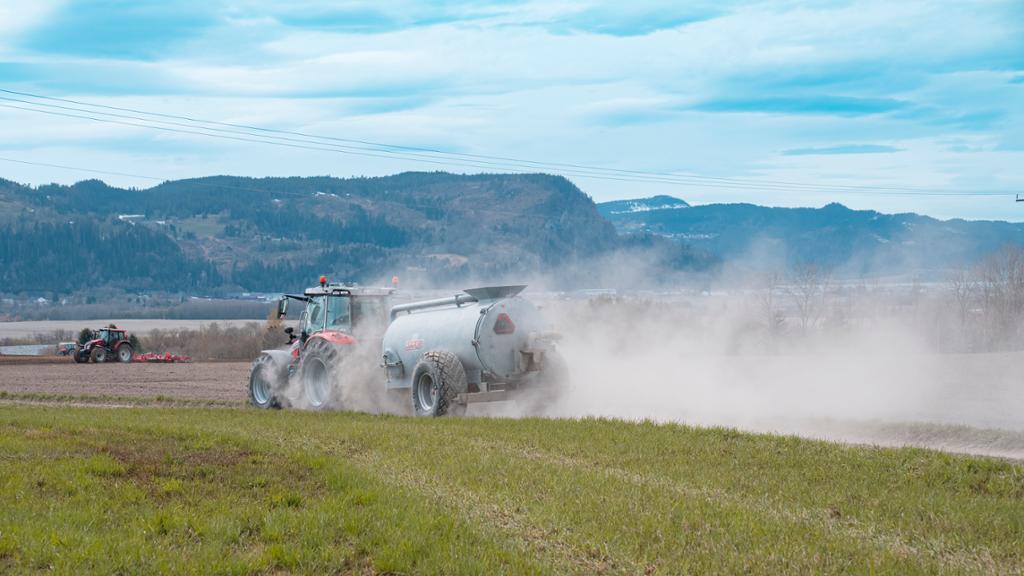 Traktor køyrer ut på eng med gjødseltankvogn. Tørr jord skaper støvsky. Foto.