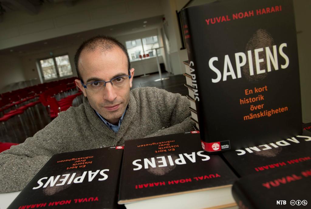 Mann står bak ei bunke med bøker. Alle bøkene har tittelen: "Sapiens". Foto.