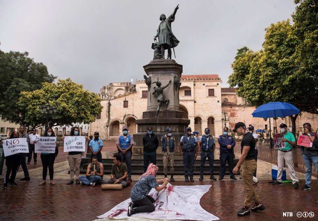 Aksjonsgruppe med plakatar framfor ein statue av ein mann i mellomalderdrakt. Foto.