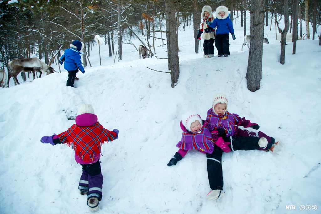 Barn leker i snøen. De har samiske sjal og luer utenpå vinterdressene. Foto. 