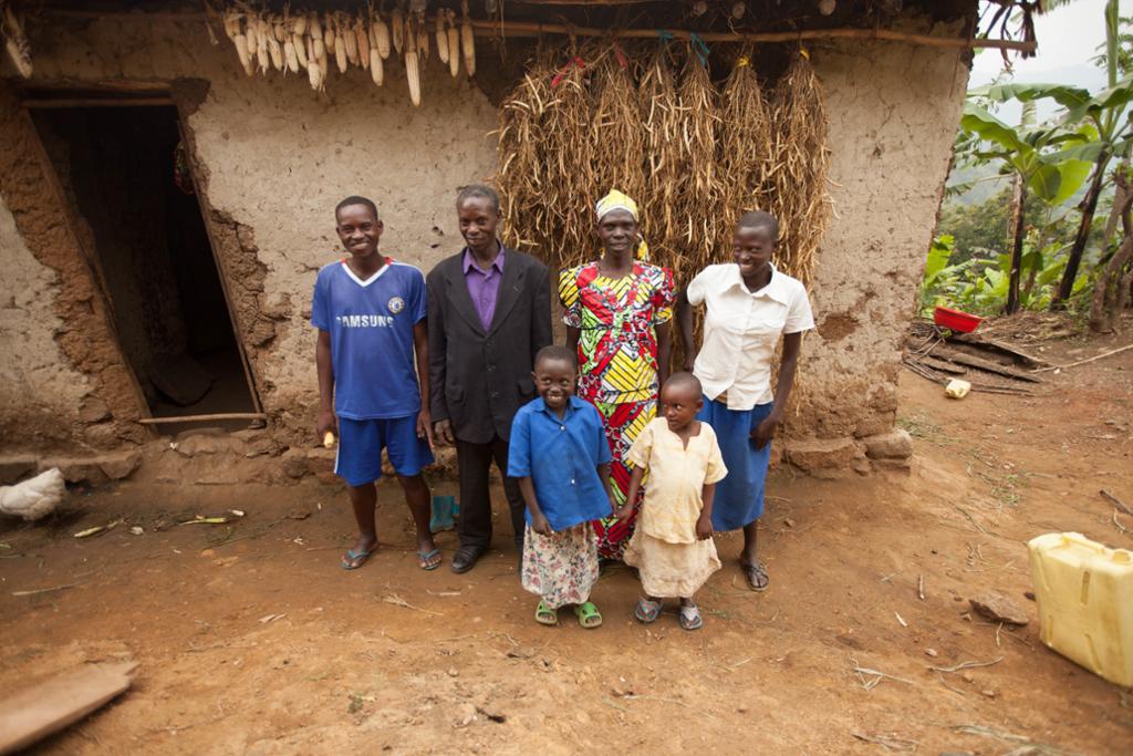 Ein smilande familie på seks personar står framfor huset sitt, som er ei bygning av sliten mur. Foto. 