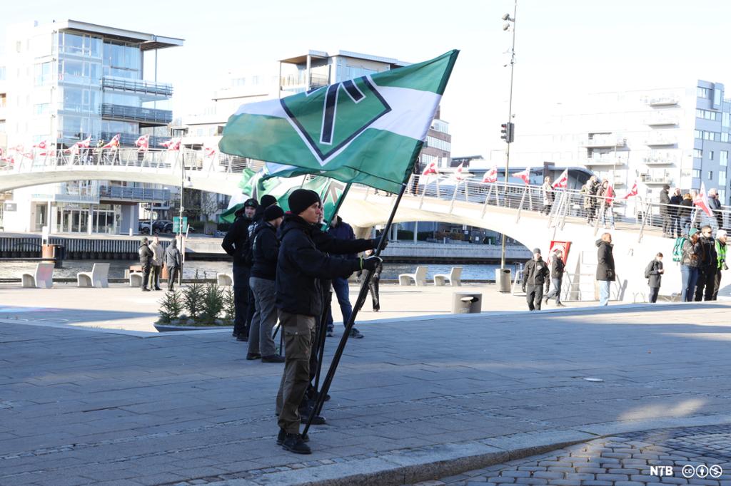 Bildet viser fem-seks unge menn  fra Den nordiske motstandsbevegelsen. De holder fram grønne flagg med runen "T". Det er noen mennesker som står på avstand rundt på plassen. Foto. 
