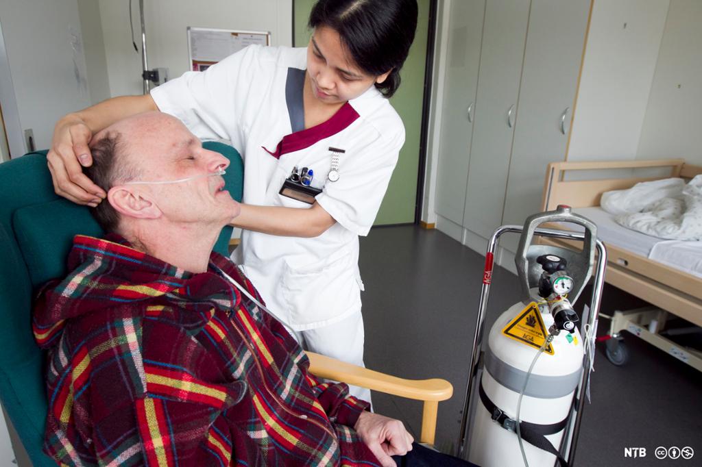 En eldre mann får ekstra oksygen gjennom et nesekateter. En kvinnelig helsefagarbeider fester oksygenslangen. Foto.