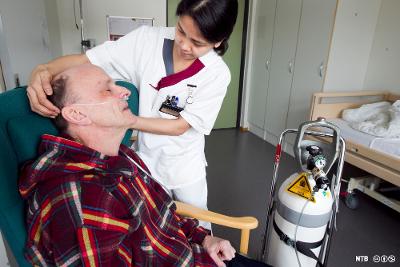 En syk eldre mann får ekstra oksygen gjennom et nesekateter . En helsefagarbeider fester oksygenslangen. Foto.