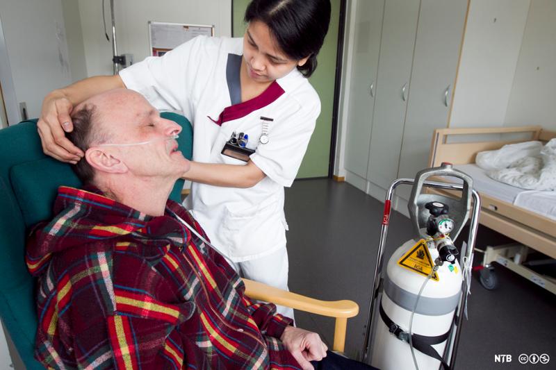 Helsefagarbeider justerer brillekateter for oksygentilførsel for mannlig pasient som sitter i stol. Foto.