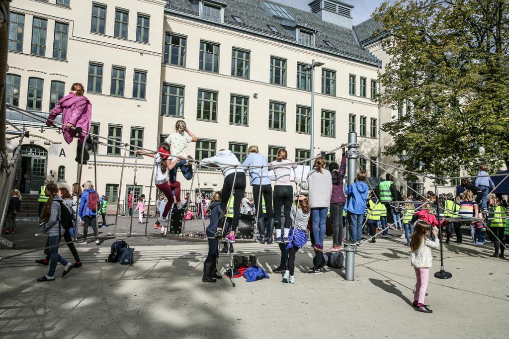 Et asfaltert uteområde foran en murbygning. Mange barn oppholder seg der, og flere klatrer og balanserer på et lekeapparat. Foto.