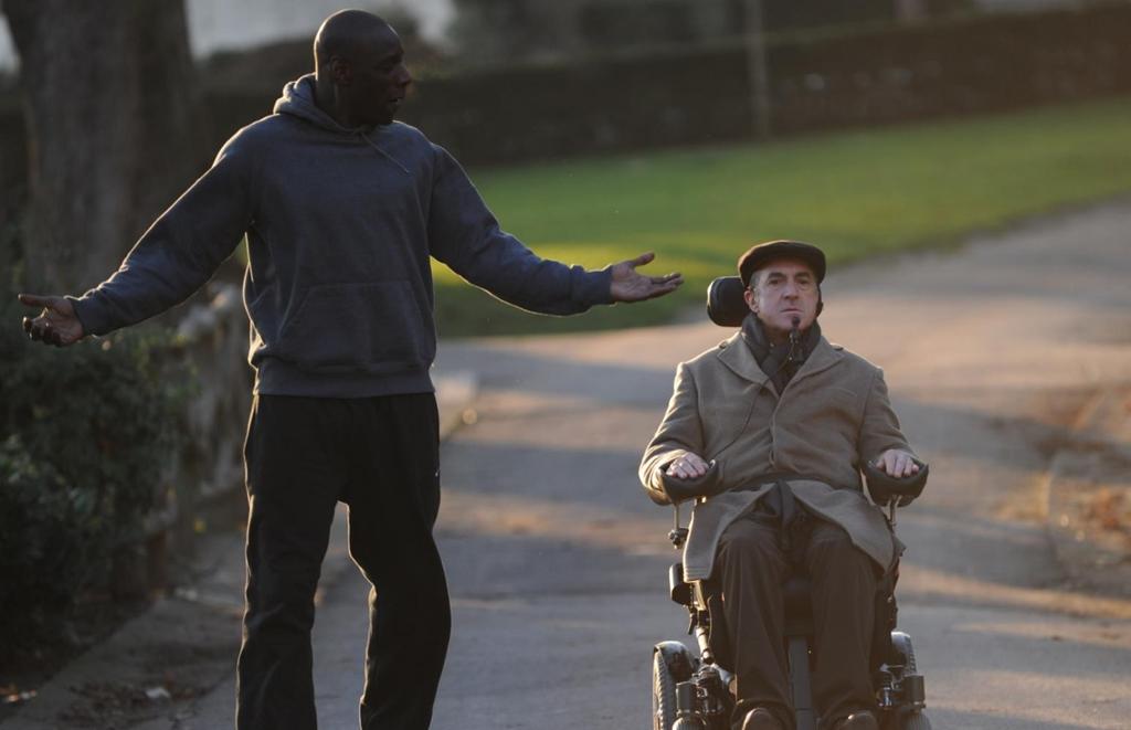 Scene fra filmen De urørlige, hvor de to hovedrolleinnehaverne er på tur i parken. Skuespiller Omar Sy i treningstøy snakker og gestikulerer, med François Cluzet i elektrisk rullestol ved siden av. Foto.