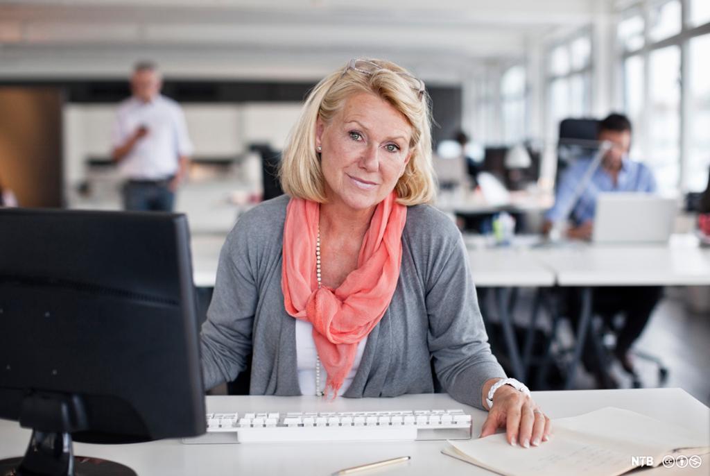 En kvinne sitter foran et arbeidsbord med dataskjerm og tastatur. I bakgrunnen skimtes flere personer. Foto.