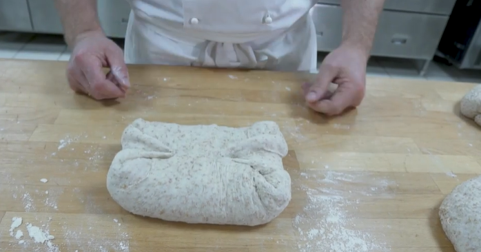 En grov brøddeig blir formet til brød på et bakebord. Foto.