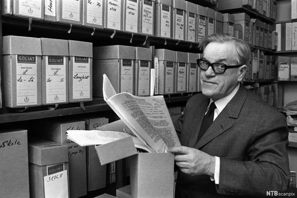 Mann med dokument framfor mange arkivboksar. Foto.