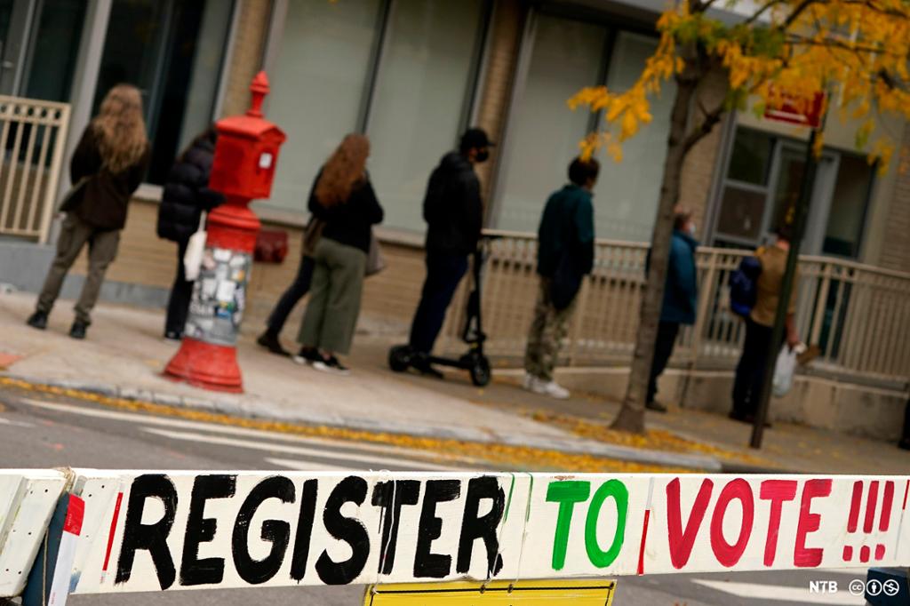 A sign urging people to register to vote is displayed near a line of people at a voting site in New York during the 2020 election. Photo.