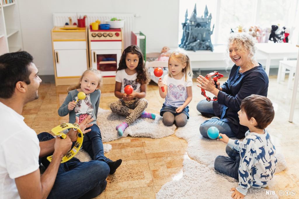 To vaksne og fire barn sit i ein ring på golvet og held ulike musikkinstrument i hendene. Foto.