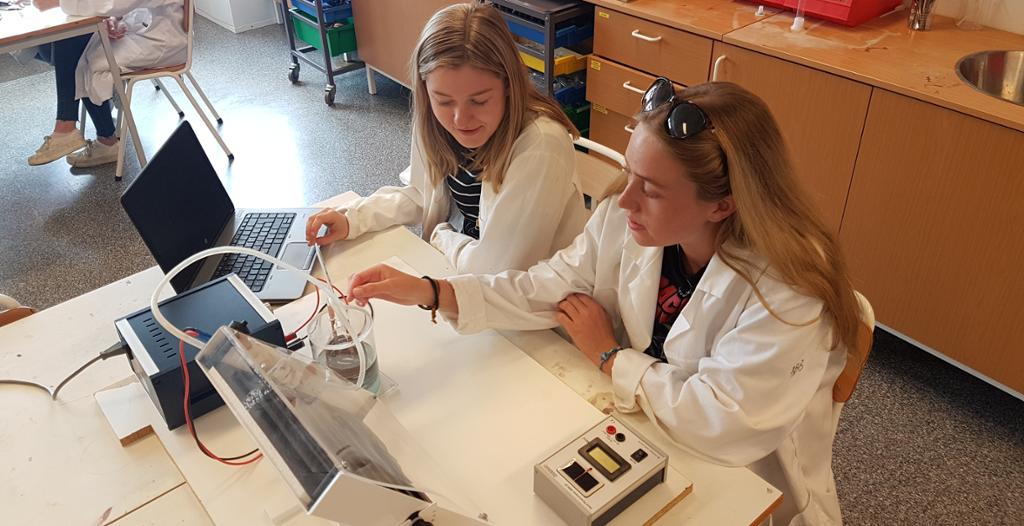 To jenter i labfrakk undersøker en modell av en solfanger. Foto.