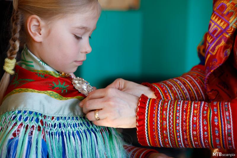 Samisk kvinne hjelper jente med å pynte seg. Vi ser jenta og armene til kvinnen som fester ei sølje til sjalet. Foto. 