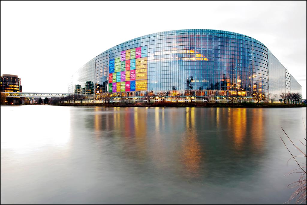 Photo: We see a modern, curved building made of steel and glass. Some of the windows appear to have posters displayed on them. The building is located by a lake which is frozen. 