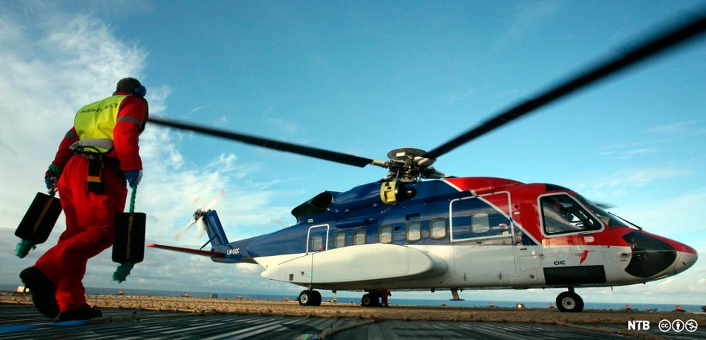 En person i kjeledress og med refleksvest og øreklokker er i på vei mot et Sikorsky S92-helikopter. Personen har en hjulkloss i hver hånd. Foto.